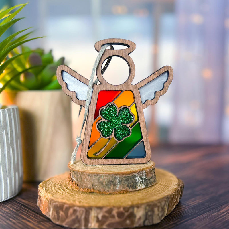 The Glittery Four-Leaf Clover Ornament, a 3.5" Mother's Angels® angel figurine by Forged Flare®, features a rainbow center and green glitter shamrock, displayed on tree slices with blurred potted plants in the background.