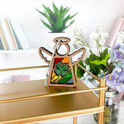A Forged Flare® 3.5" Mother's Angels® figurine stands on a gold shelf with a rainbow backdrop and a glittery four-leaf clover ornament. Behind are books, a green plant, and vases with white and purple flowers.