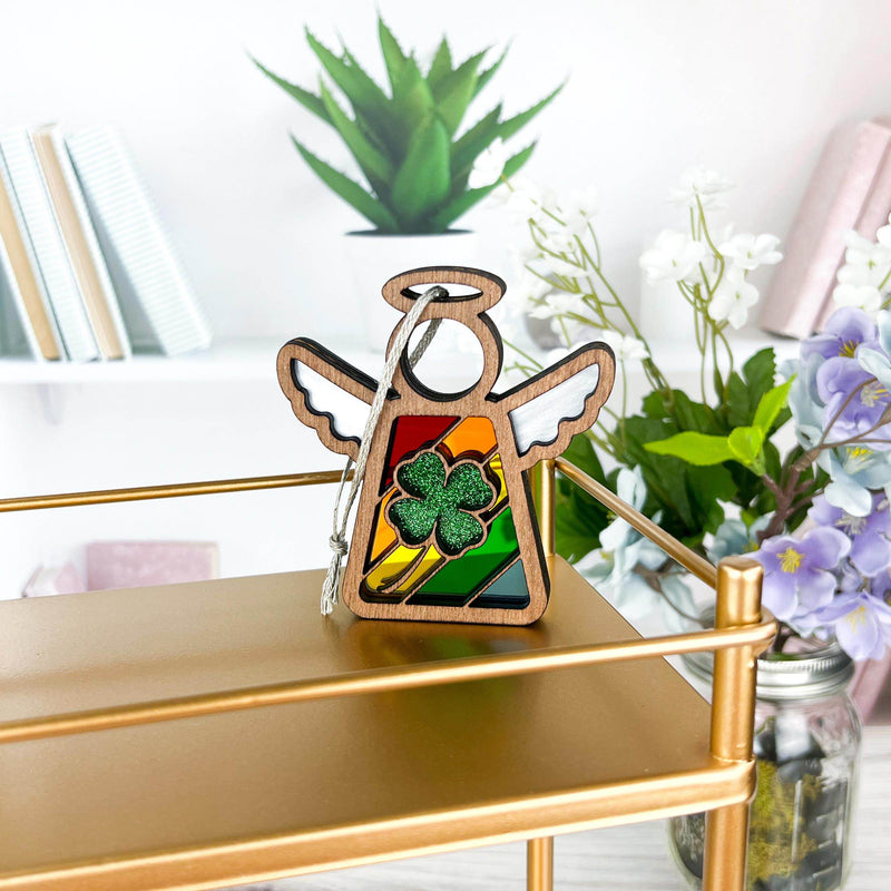 A Forged Flare® 3.5" Mother's Angels® figurine stands on a gold shelf with a rainbow backdrop and a glittery four-leaf clover ornament. Behind are books, a green plant, and vases with white and purple flowers.