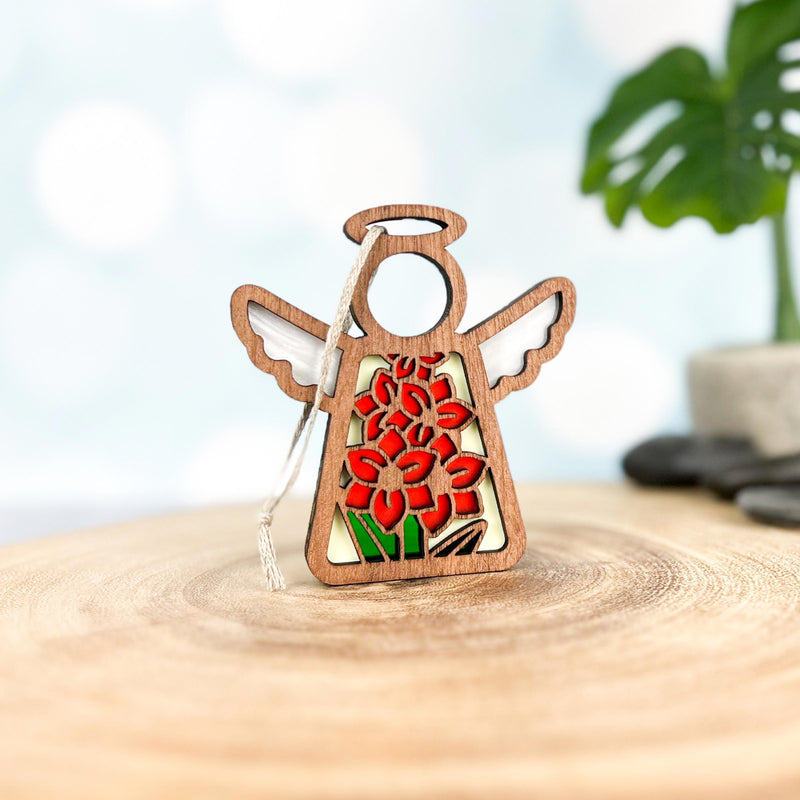 The Forged Flare® Gladiolus Ornament | 3.5" (H) Mother's Angels® features a wooden angel figurine with a red gladiolus flower design, white hanging string, and is displayed on a round wooden surface with green plants and gray stones in the background.