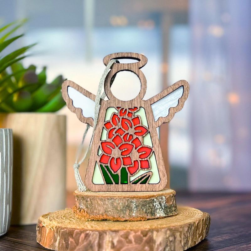 The Forged Flare® Gladiolus Ornament, a 3.5" Mother's Angels® figurine with white wings and stained-glass-style red gladiolus (August birth flower), stands on a tree slice beside a green plant against a softly lit background.