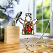 The Forged Flare® Gladiolus Ornament | 3.5" (H) Angel Figurine from the Mother's Angels® collection hangs on a desk stand, with blurred flowers, a wooden pen holder, and sunlight streaming through the window in the background.