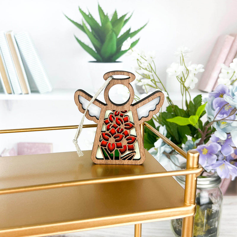 A Forged Flare® Gladiolus Ornament | 3.5" (H) Angel Figurine from the Mother's Angels® line stands on a gold shelf, with books, August birth flower gladiolus, white and purple blooms, and green plants in the background.