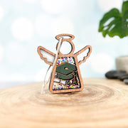 The Forged Flare® Mother's Angels® 3.5" Graduation Ornament features a wooden angel with halo and wings, graduation cap, diploma, and colorful dots—perfect for graduations, displayed on a wood surface with a plant in the background.