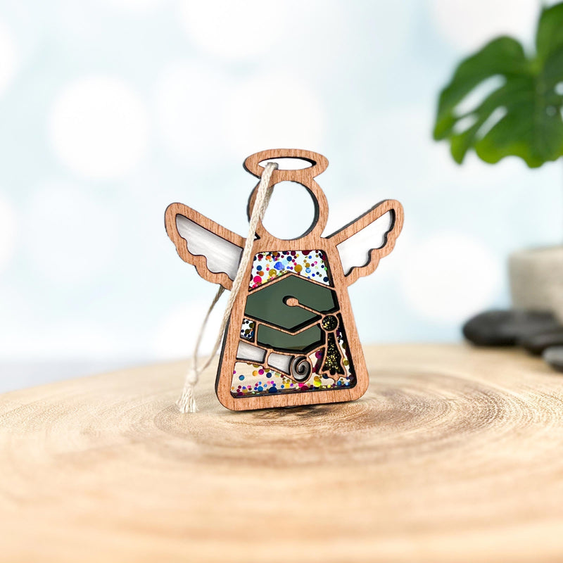 The Forged Flare® Mother's Angels® 3.5" Graduation Ornament features a wooden angel with halo and wings, graduation cap, diploma, and colorful dots—perfect for graduations, displayed on a wood surface with a plant in the background.