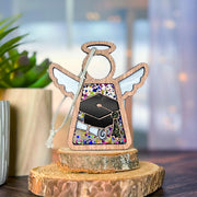 The Forged Flare® Mother's Angels® Graduation Ornament is a 3.5" wooden angel figurine with white wings, a robe adorned with a graduation cap and diploma, standing on wood slices—perfect for celebrating academic achievement.