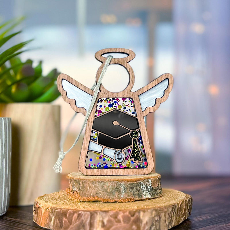 The Forged Flare® Mother's Angels® Graduation Ornament is a 3.5" wooden angel figurine with white wings, a robe adorned with a graduation cap and diploma, standing on wood slices—perfect for celebrating academic achievement.