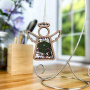 The Forged Flare® Mother's Angels® Graduation Ornament, a 3.5" wooden angel figurine with stained glass, features a green graduation cap and diploma, making it a thoughtful gift. It hangs from a stand on a wooden desk with blurred flowers in the background.