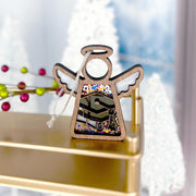 The Forged Flare® Mother's Angels® Graduation Ornament is a 3.5" angel figurine with painted wings and colorful patterns, displayed on a gold shelf, perfect for graduation celebrations amid festive Christmas trees and red berries in the background.