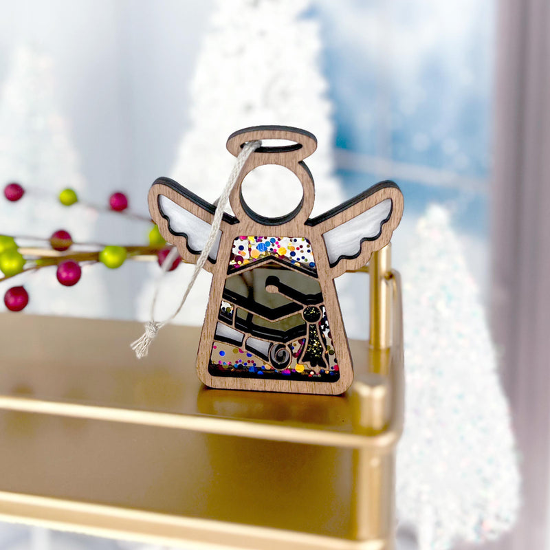 The Forged Flare® Mother's Angels® Graduation Ornament is a 3.5" angel figurine with painted wings and colorful patterns, displayed on a gold shelf, perfect for graduation celebrations amid festive Christmas trees and red berries in the background.