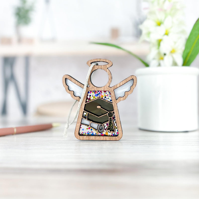 A charming Graduation Ornament from Forged Flare's® Mother's Angels® collection stands on a light-colored table alongside a white potted plant. This 3.5" angel figurine, adorned with a colorful, patterned torso and a graduation cap, is the perfect addition to any angel figurines collection or as unique graduation party decor.