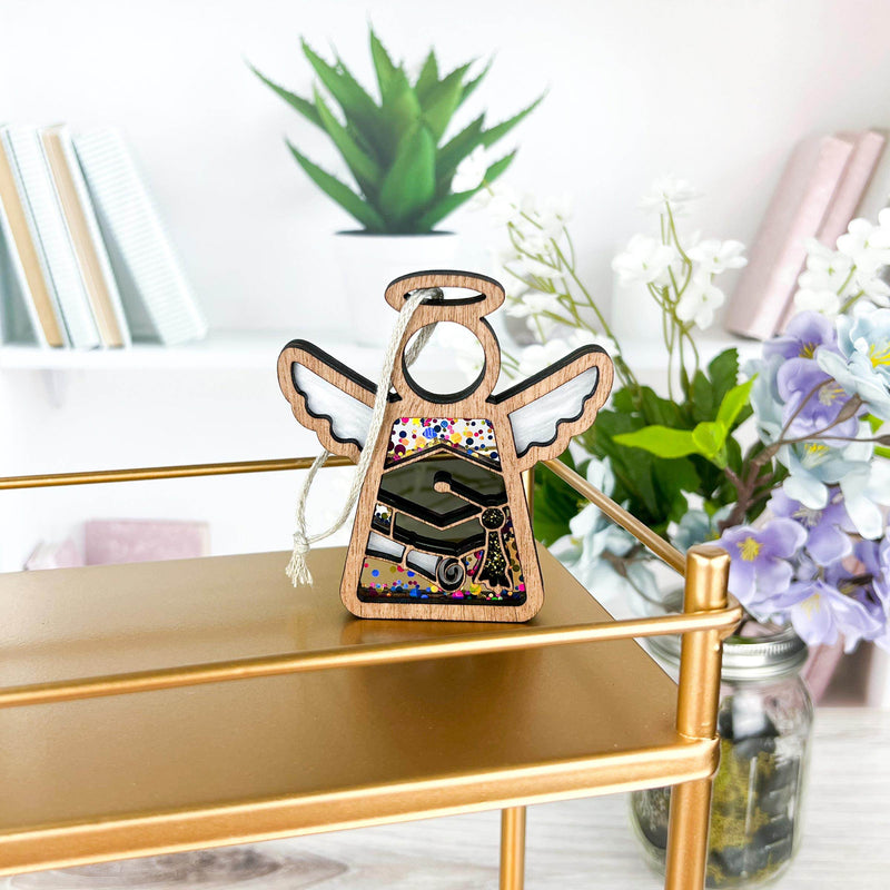 The Forged Flare® Mother's Angels® Graduation Ornament, a 3.5" angel figurine with white wings and a colorful dress, stands on a gold shelf amid books, a potted plant, and flowers for a bright, stylish display.