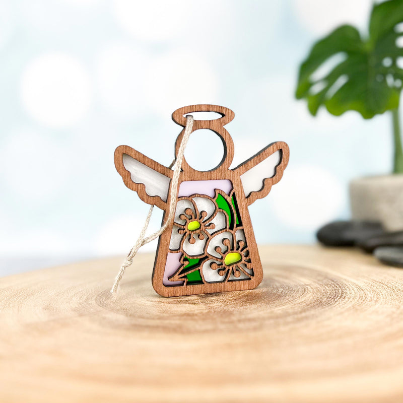 The Forged Flare® Hawthorn Ornament is a 3.5" Mother's Angels® wooden angel figurine, featuring delicate hawthorn flower cutouts and colored accents, with a string halo. A potted plant is softly blurred in the background.