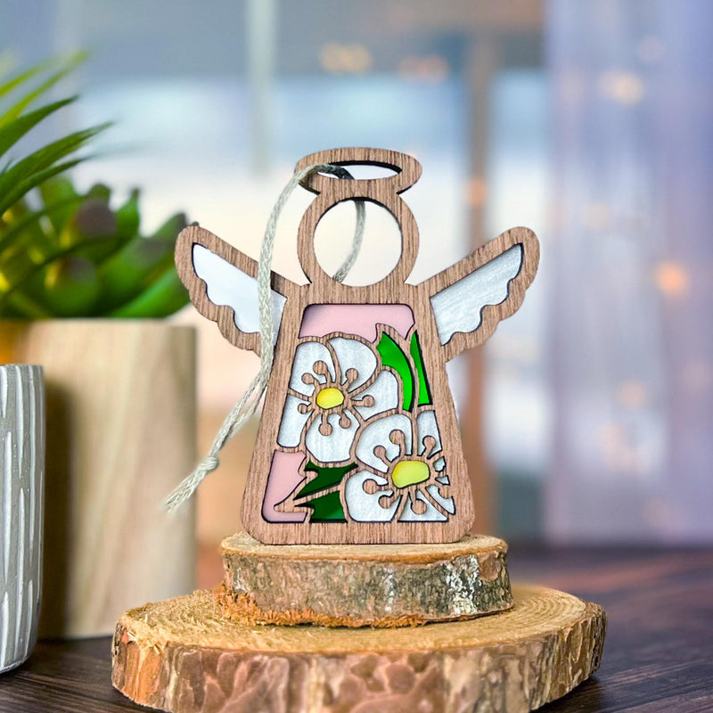 The Forged Flare® Hawthorn Ornament | 3.5" (H) Angel Figurine from the Mother's Angels® collection stands among tree slice coasters, adorned with delicate hawthorn flowers and white wings in a cozy indoor setting with a potted plant nearby.