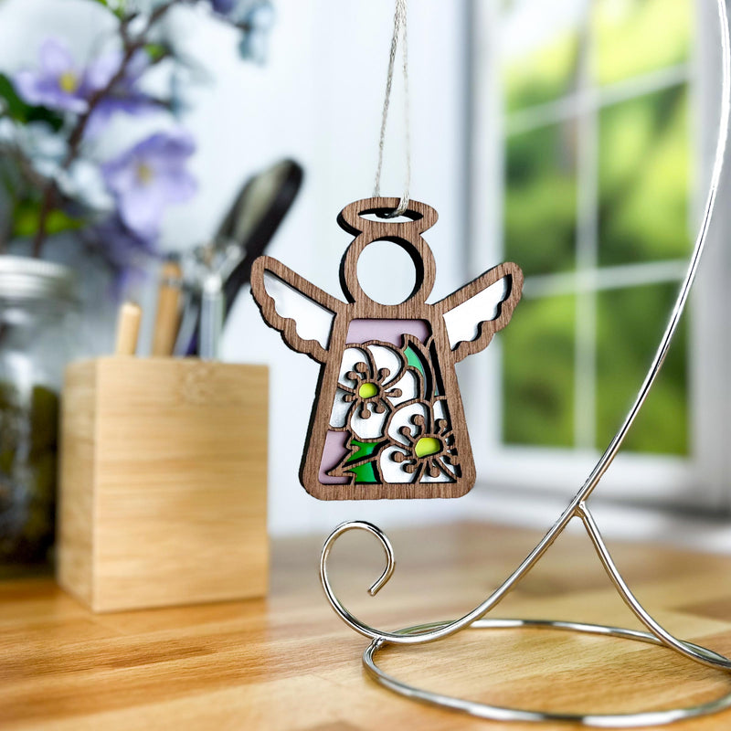The Forged Flare® Mother's Angels® Hawthorn Ornament, a 3.5" wooden angel figurine with hawthorn flower cutouts, hangs from a stand on a table, with a blurred window, flowers, and utensils in the background.