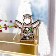 The Forged Flare® Hawthorn Ornament | 3.5" (H) Angel Figurine from the Mother's Angels® line features delicate flower cutouts and white wings, displayed on a gold shelf with blurred white Christmas trees and red berries in the background.