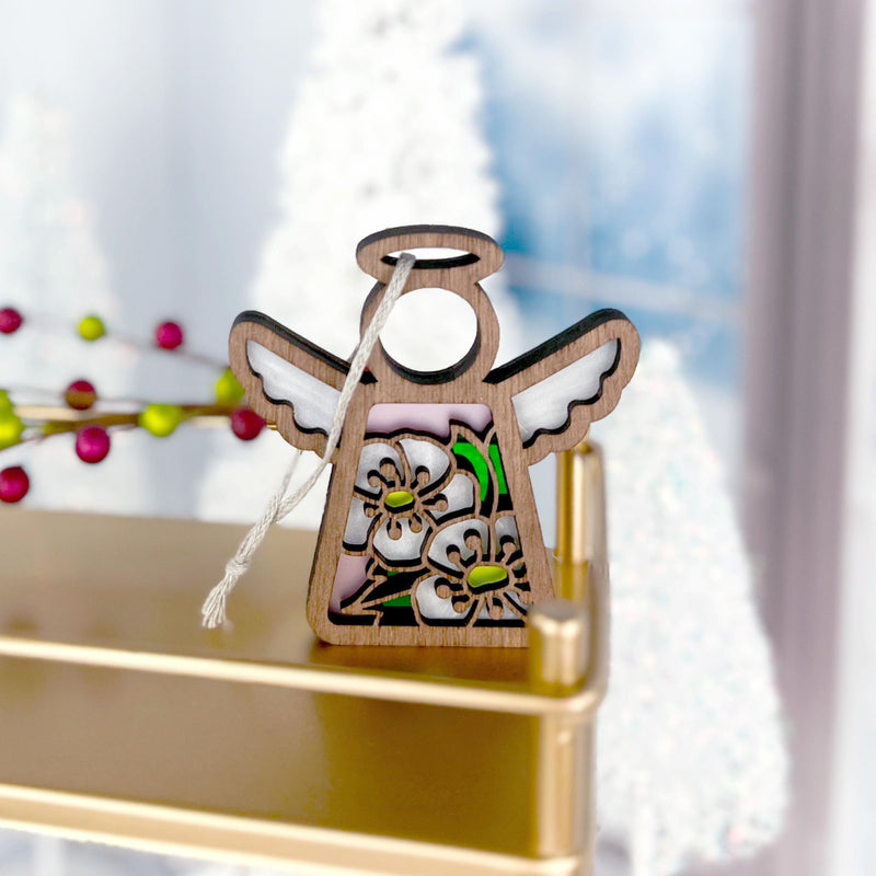 The Forged Flare® Hawthorn Ornament | 3.5" (H) Angel Figurine from the Mother's Angels® line features delicate flower cutouts and white wings, displayed on a gold shelf with blurred white Christmas trees and red berries in the background.