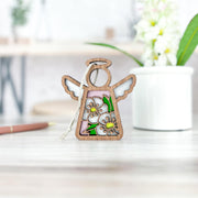 The Forged Flare® Hawthorn Ornament, a 3.5" Mother's Angels® wooden angel adorned with painted white hawthorn flowers and green leaves, stands on a light table with a blurred potted plant and brown pen in the background.