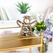 The Forged Flare® Hawthorn Ornament, a 3.5" Mother's Angels® angel figurine with floral designs, stands on a gold shelf with potted plants, books, and a jar in the background among purple and white flowers.