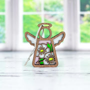 The Forged Flare® Hawthorn Ornament is a 3.5" wooden Mother's Angels® figurine with white wings and two painted hawthorn flowers, pictured on a white surface with blurred windows and greenery in the background.