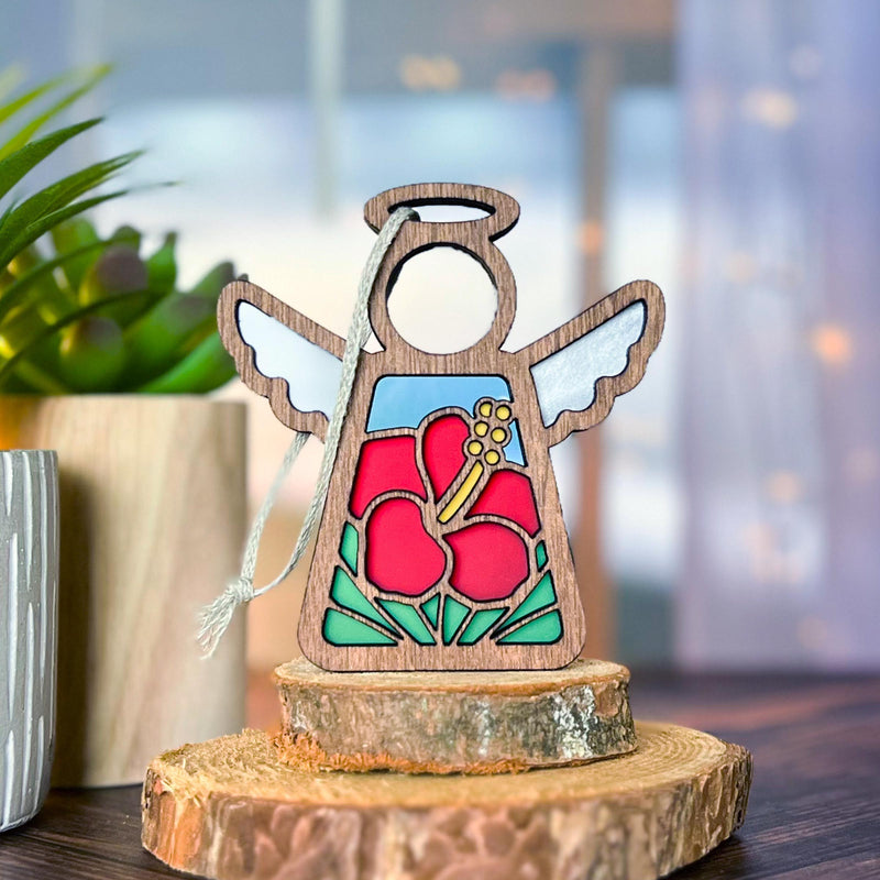 A Forged Flare® Hibiscus Ornament—a 3.5" Mother's Angels® wooden angel figurine with white wings and red hibiscus design—rests on tree slice coasters, with a green plant and soft lighting adding festive warmth.