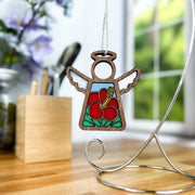 The Hibiscus Ornament, a 3.5" angel figurine from the Mother's Angels® collection by Forged Flare®, features colorful stained glass wings and a flower design in the center. It hangs gracefully from a metal stand on a wooden table. In the background, blurred flowers, a container with pens, and a window showcasing green foliage can be seen.