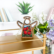 A Hibiscus Ornament 3.5" Angel Figurine from the Mother's Angels® collection by Forged Flare®, adorned with decorative red flowers and green leaves painted on its wooden body, is displayed on a golden shelf. In the background, there are potted plants, white and purple flowers, and a white shelf with books.