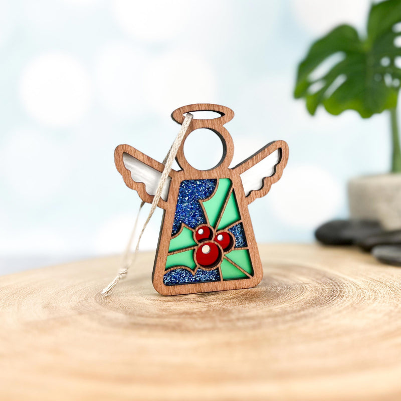 The Forged Flare® Holly Ornament | 3.5" (H) Angel Figurine from Mother's Angels® features white wings, a halo, and a glittery blue body with holly flower and red berry accents, displayed on wood with a blurred plant in the background.