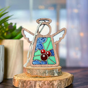 The Forged Flare® Holly Ornament | 3.5" (H) Angel Figurine from Mother's Angels® features blue and green stained glass, red holly berries, and white wings on a wooden base—evoking December birth flowers beside a softly blurred potted plant.