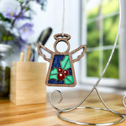 The Forged Flare® Holly Ornament is a 3.5" (H) Mother's Angels® figurine with green, red, and blue stained glass-style details. Hanging from a stand on a desk near a window, blurred holly flowers appear in the background.