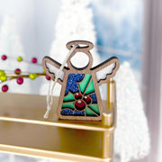 The Forged Flare® Holly Ornament is a 3.5" Mother's Angels® figurine with blue glitter wings and a green holly-inspired body, shown on a gold surface with blurred white Christmas trees in the background.