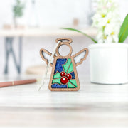 The Forged Flare® Holly Ornament | 3.5" (H) Angel Figurine from Mother's Angels® rests on a light wooden surface. A softly blurred white mug with holly flowers and a pen appear in the background.