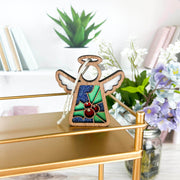 The Forged Flare® Holly Ornament | 3.5" (H) Angel Figurine from Mother's Angels® features white wings and a holly stained glass design, shown on a gold shelf with books and white and purple flowers in the background.