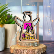 The Forged Flare® Warehouse Return - Honey Bee Ornament (3.5" Mother's Angels® angel figurine) features a stained glass-style bee and pink background, displayed with wood slices, a potted plant, and cozy lighting for an inviting look.