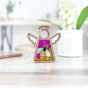 The Forged Flare® Warehouse Return - Honey Bee Ornament, a 3.5" Mother's Angels® angel figurine, features purple and yellow stained glass and wood. Displayed on a light wooden table with blurred flowers and a white pot in the background. Available on Amazon.