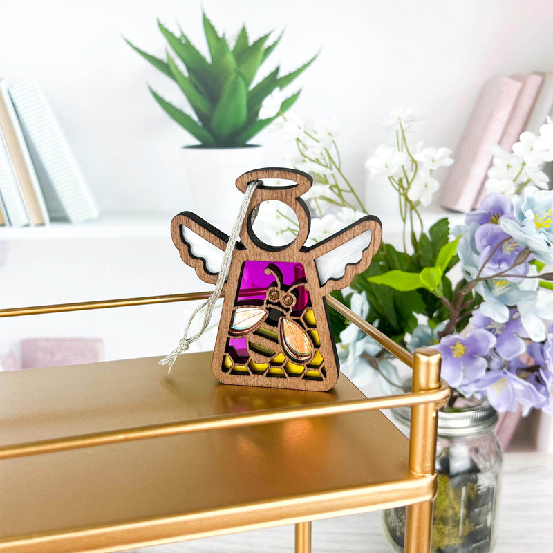 A 3.5" Honey Bee Mother's Angels® ornament by Forged Flare® with a stained glass-style design in purple, pink, and yellow stands on a gold shelf. Books, a potted plant, and a vase with white and purple flowers are in the background.