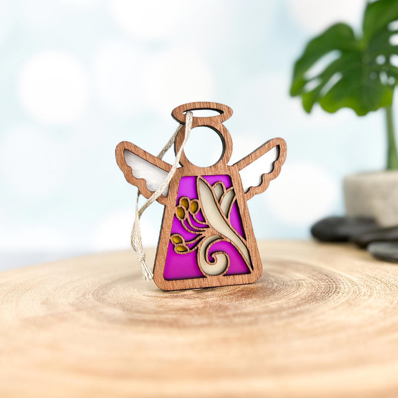 The Honeysuckle Ornament | 3.5" (H) Angel Figurine by Forged Flare® features intricate cut-outs, gold swirls, and June’s honeysuckle flower on wood with a purple background; blurred plants and stones appear behind.