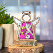 The Forged Flare® Honeysuckle Ornament is a 3.5" Mother's Angels® figurine with white wings and birth flower art on purple, displayed on a wood slice beside a potted plant, with a softly blurred background.