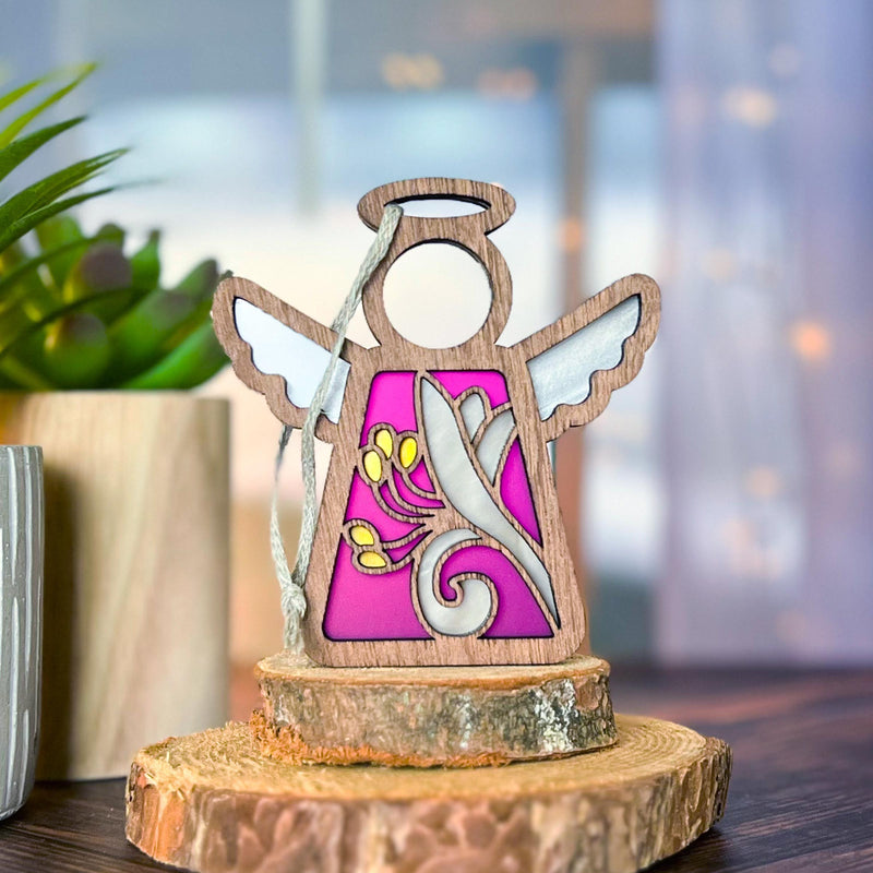 The Forged Flare® Honeysuckle Ornament is a 3.5" Mother's Angels® figurine with white wings and birth flower art on purple, displayed on a wood slice beside a potted plant, with a softly blurred background.