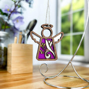 A Forged Flare® Honeysuckle Ornament, a 3.5" Mother's Angels® angel figurine with a purple and gold swirl, hangs from a curved metal stand on a desk with blurred flowers, pencils, and a window in the background.