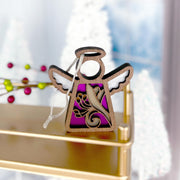 The Forged Flare® Honeysuckle Ornament, a 3.5" Mother's Angels® figurine with white wings and a honeysuckle flower design on purple, rests on a golden shelf with blurred Christmas trees and berries in the background.