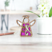 The 3.5" Honeysuckle Ornament angel figurine by Forged Flare® Mother's Angels® features intricate floral cutouts and sits on a light wooden surface with a purple background; a white potted plant and pen blur softly behind.