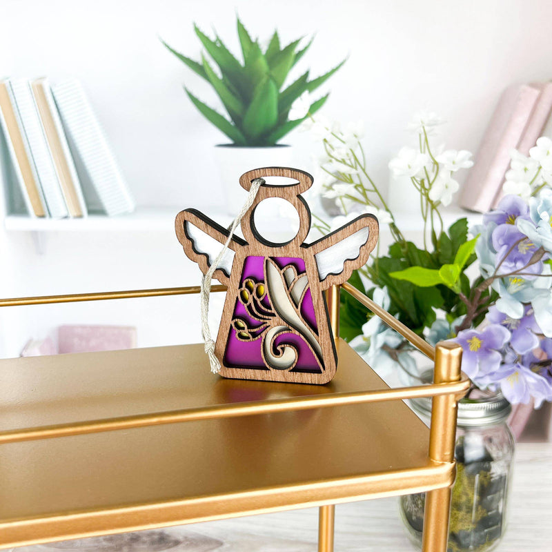 A Forged Flare® Honeysuckle Ornament | 3.5" (H) Mother's Angels® figurine with white wings and gold accents stands on a gold shelf, surrounded by books, a green potted plant, and a jar with purple and honeysuckle flowers.