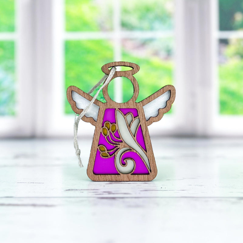 The Forged Flare® Honeysuckle Ornament is a 3.5" Mother's Angels® wooden angel figurine with white wings, a pink and gold honeysuckle flower pattern, and a hanging string, shown on a white surface with a blurred green window background.