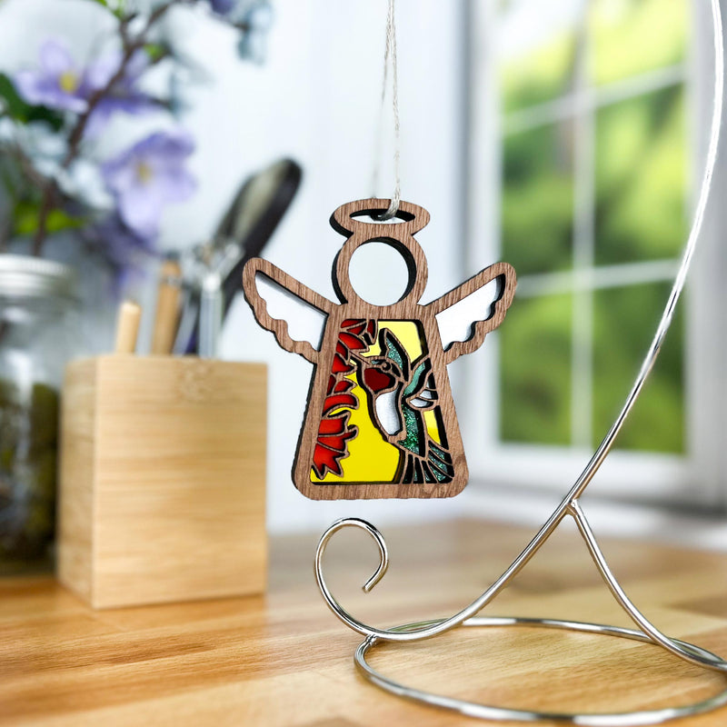 A Forged Flare® Mother's Angels® Ruby-Throated Hummingbird Ornament stands on a metal base atop a wooden desk by the window, its stained glass-style wings and bird design bright against softly blurred greenery and purple flowers.