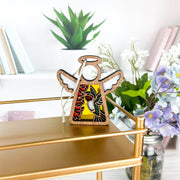 A Forged Flare® Mother's Angels® 3.5" Ruby-Throated Hummingbird ornament, featuring stained glass wings and floral accents, hangs on a gold shelf with books, a green plant, and purple-white flowers in the background.