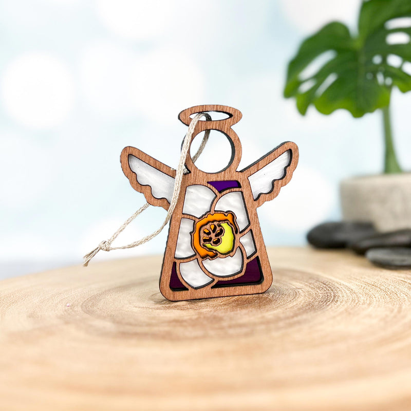 The Forged Flare® Jonquil Ornament, a 3.5" Mother's Angels® wooden angel with white wings and a stained glass-style March birth flower, stands on a wooden surface with a blurred plant in the background.