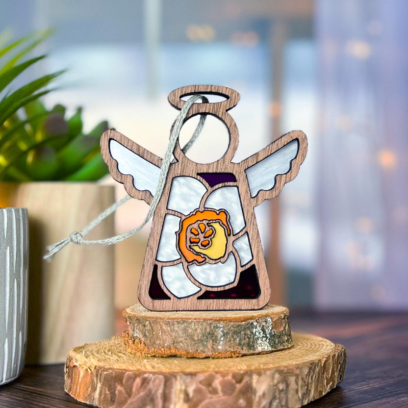 The Forged Flare® Jonquil Ornament, a 3.5" Mother's Angels® angel figurine with white wings and jonquil flower design, stands upright on stacked tree slices, framed by a potted plant and soft-focus background.