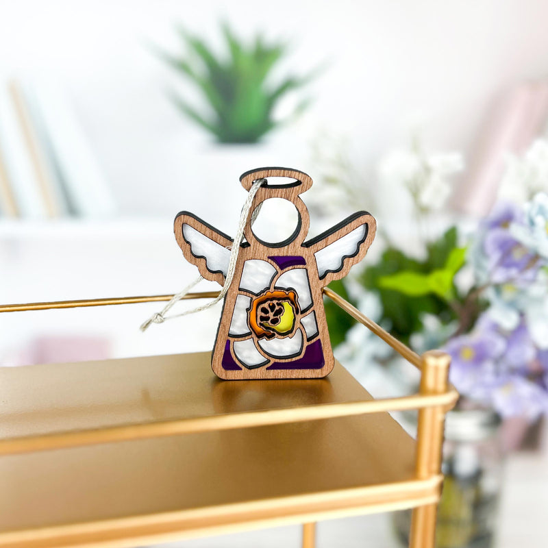 The Forged Flare® Mother's Angels® Jonquil Ornament, a 3.5" wooden angel figurine with white wings and a jonquil flower for March, sits on a gold shelf with blurred plants and books in the background.