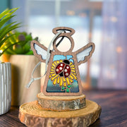 The Ladybug and Dandelion Ornament by Forged Flare® is a 3.5" wooden angel figurine featuring stained-glass-style art, perfect as a Mother's Angels® Christmas tree ornament or gift, shown on wood slices with plants and a blurred window behind.